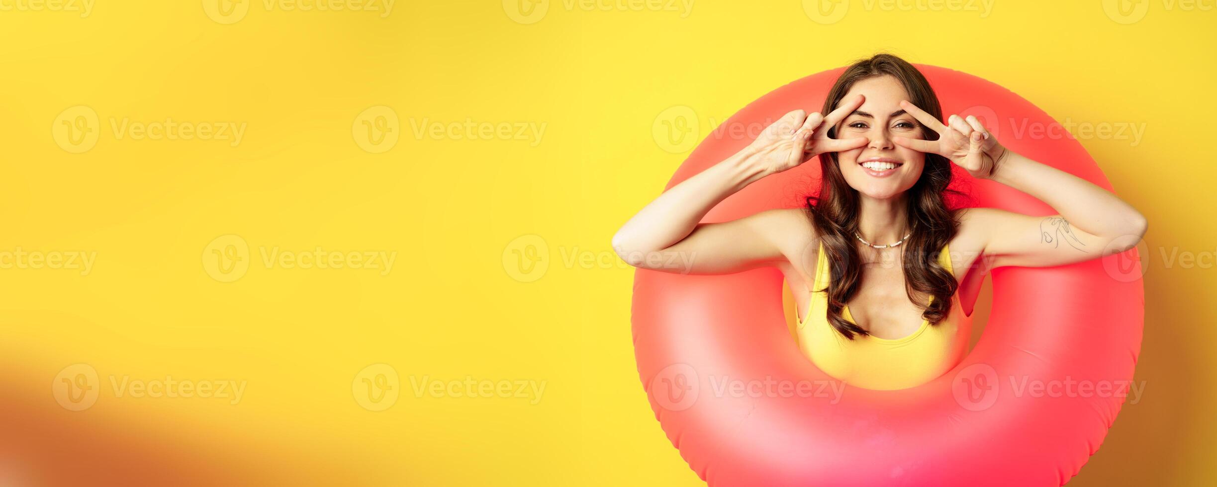 Portrait of attractive modern girl inside trendy pink beach swimming ring, showing peace v-sign and smiling happy, standing over yellow background photo