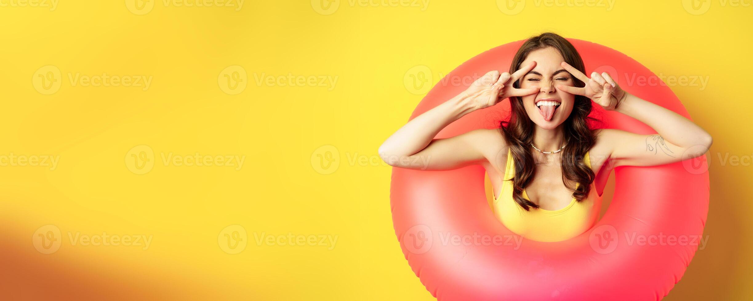 Portrait of attractive modern girl inside trendy pink beach swimming ring, showing peace v-sign and smiling happy, standing over yellow background photo