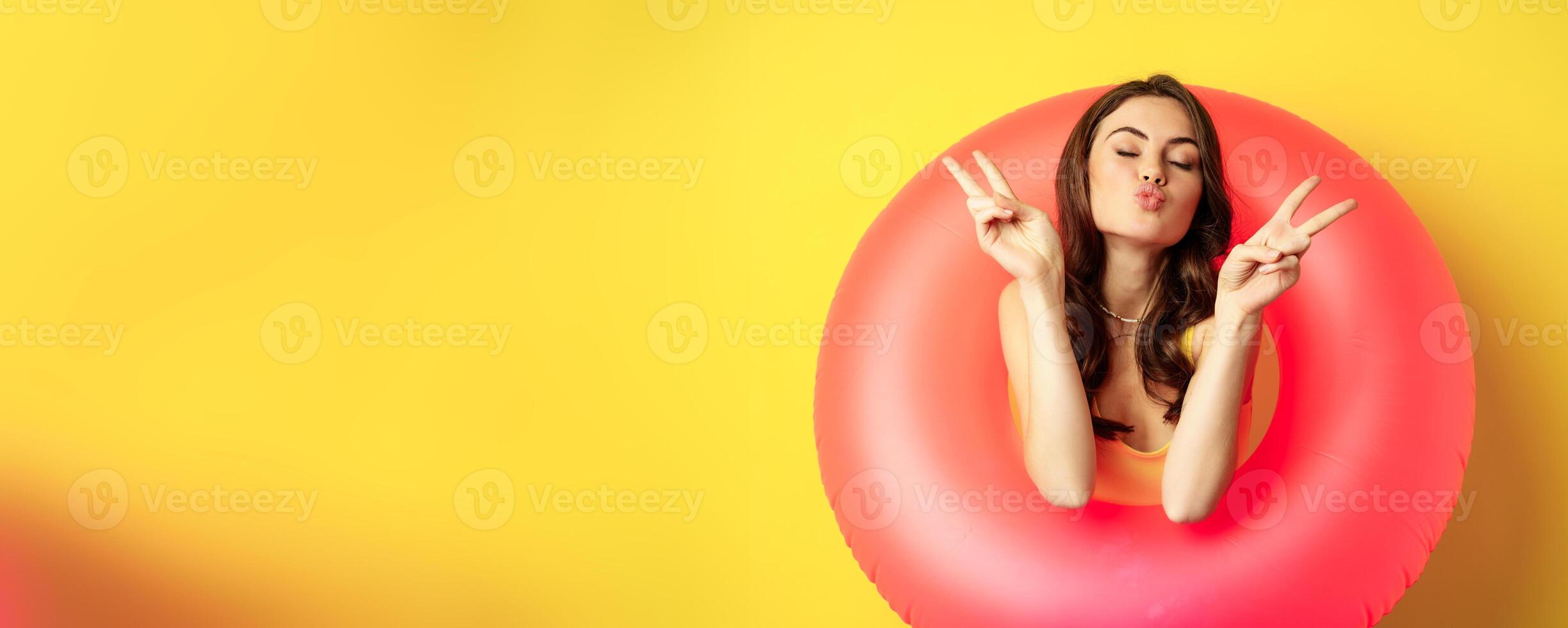 Portrait of attractive modern girl inside trendy pink beach swimming ring, showing peace v-sign and smiling happy, standing over yellow background photo