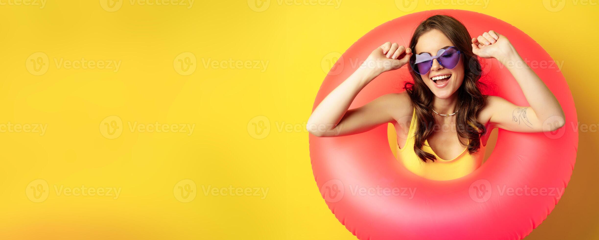 Close up portrait of stylish young woman, girl with swimming ring and summer sunglasses, ready for beach holiday, vacation, standing over yellow background photo