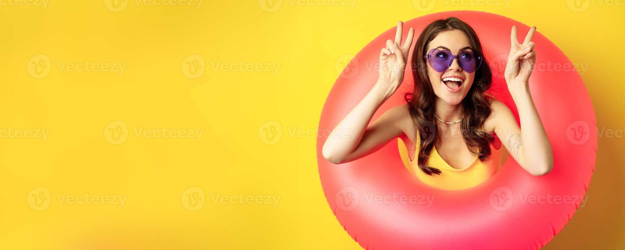 Portrait of happy stylish girl in sunglasses, inside beach swimming ring, showing peace sign, laughing and smiling, standing over yellow background photo