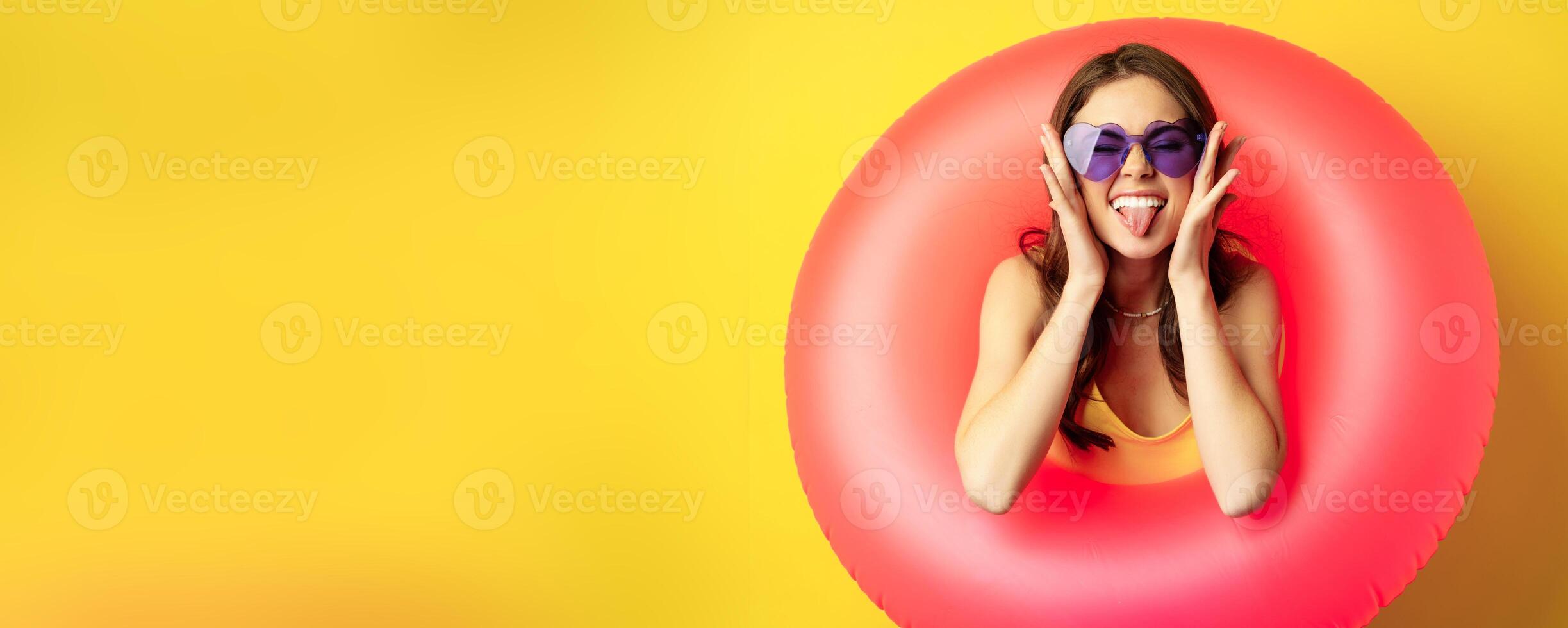 Close up portrait of enthusiastic young woman inside pink swimming ring, laughing and smiling, enjoying beach holiday, summer vacation, yellow background photo