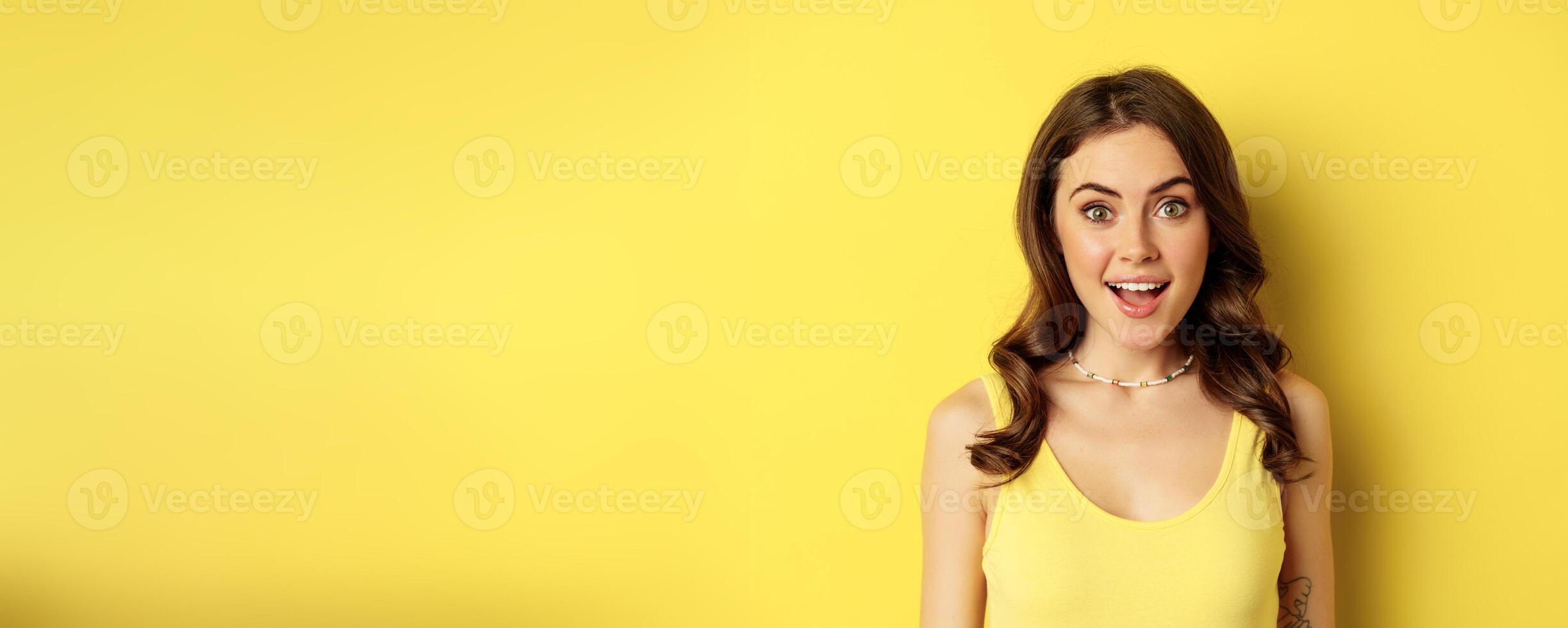 Close up portrait of stylish brunette girl looking amazed, surprised face expression, saying wow, reacting impressed, standing over yellow background photo
