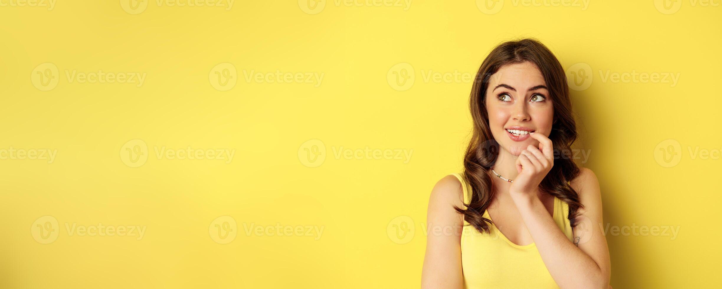 Close up portrait of stylish young beautiful woman thinking, yearning smth, looking up and making choice, standing over yellow background photo
