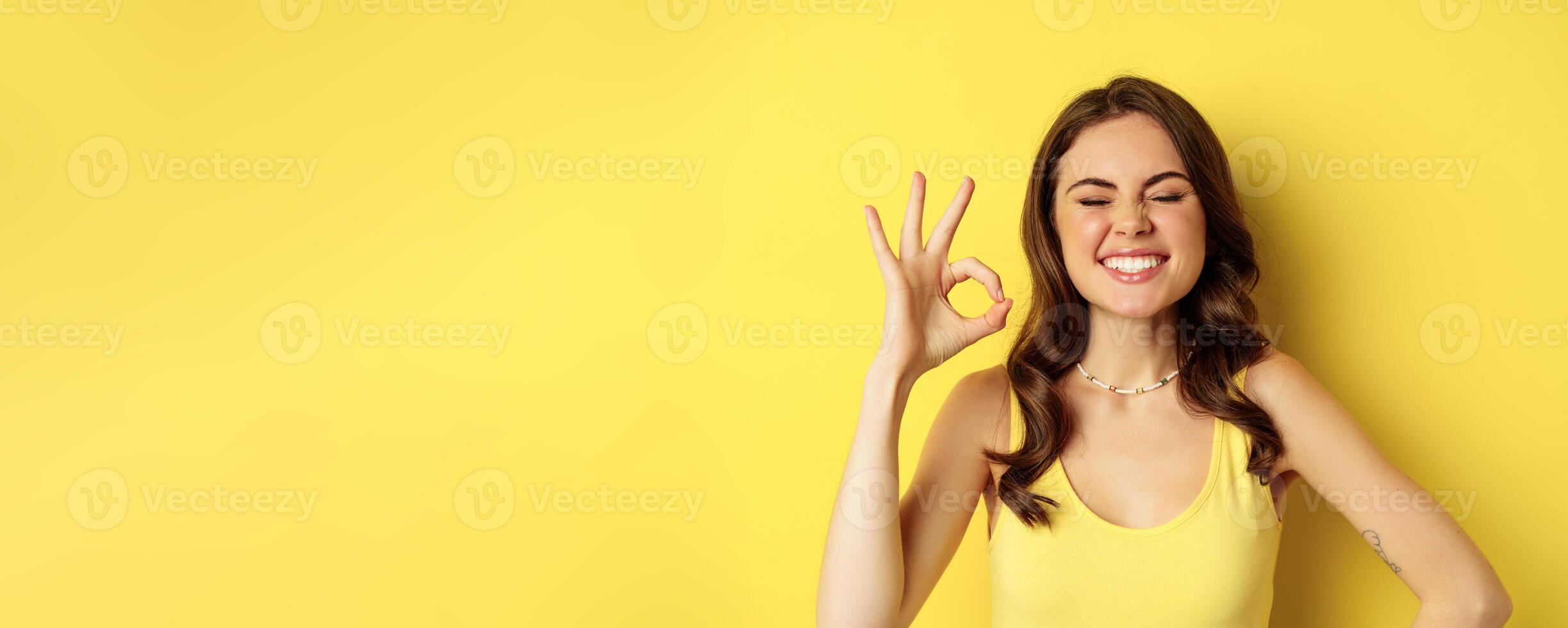 Close up portrait of young happy pretty woman, laughing and smiling with white teeth, showing excellent, okay ok sign, praise and compliment smth, yellow background photo