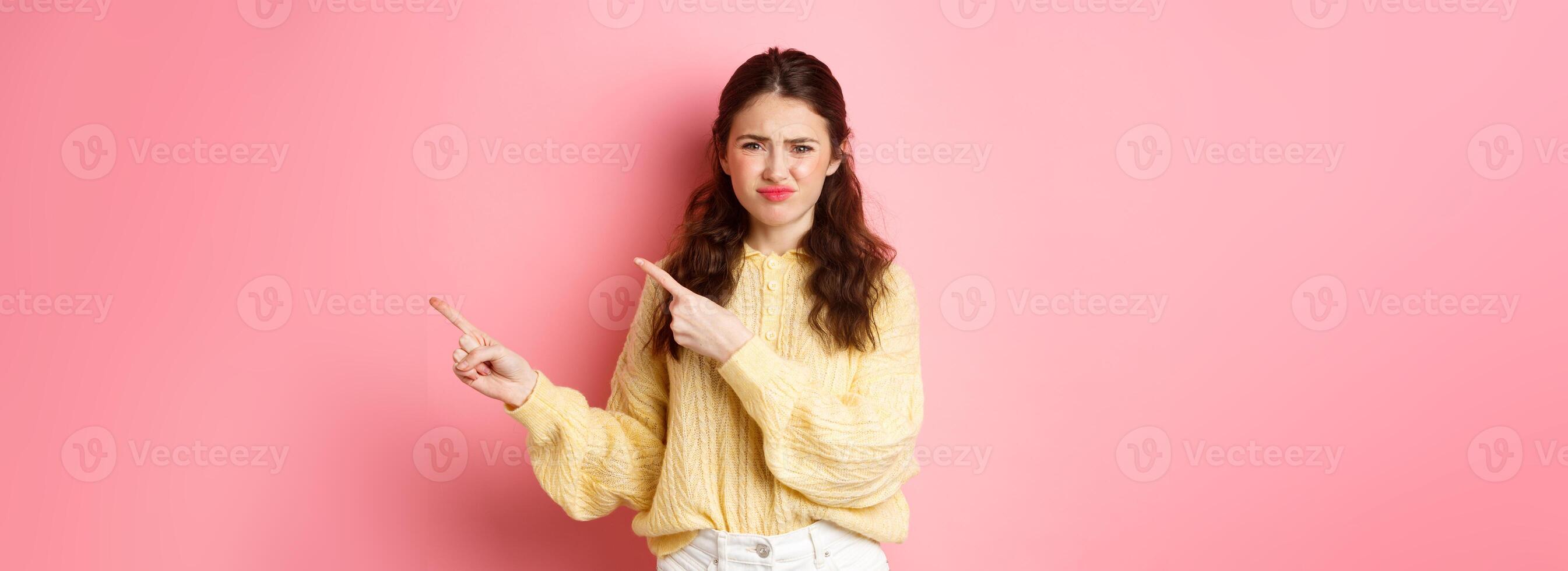Skeptical and displeased young woman frowning, grimacing and pointing fingers right at copyspace, express dislike and disappointment, standing against pink background photo