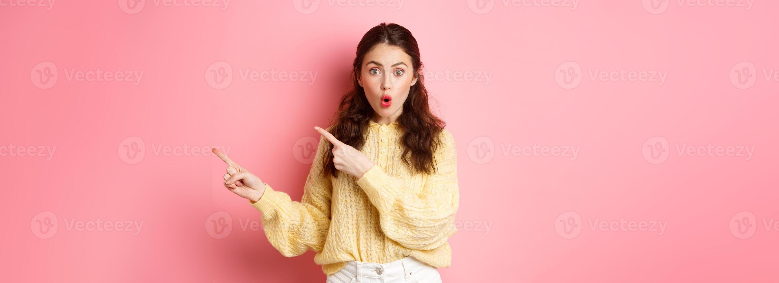 Portrait of surprised brunette girl gasping impressed, pointing fingers right at copy space aside, showing way to logo, standing over pink background photo