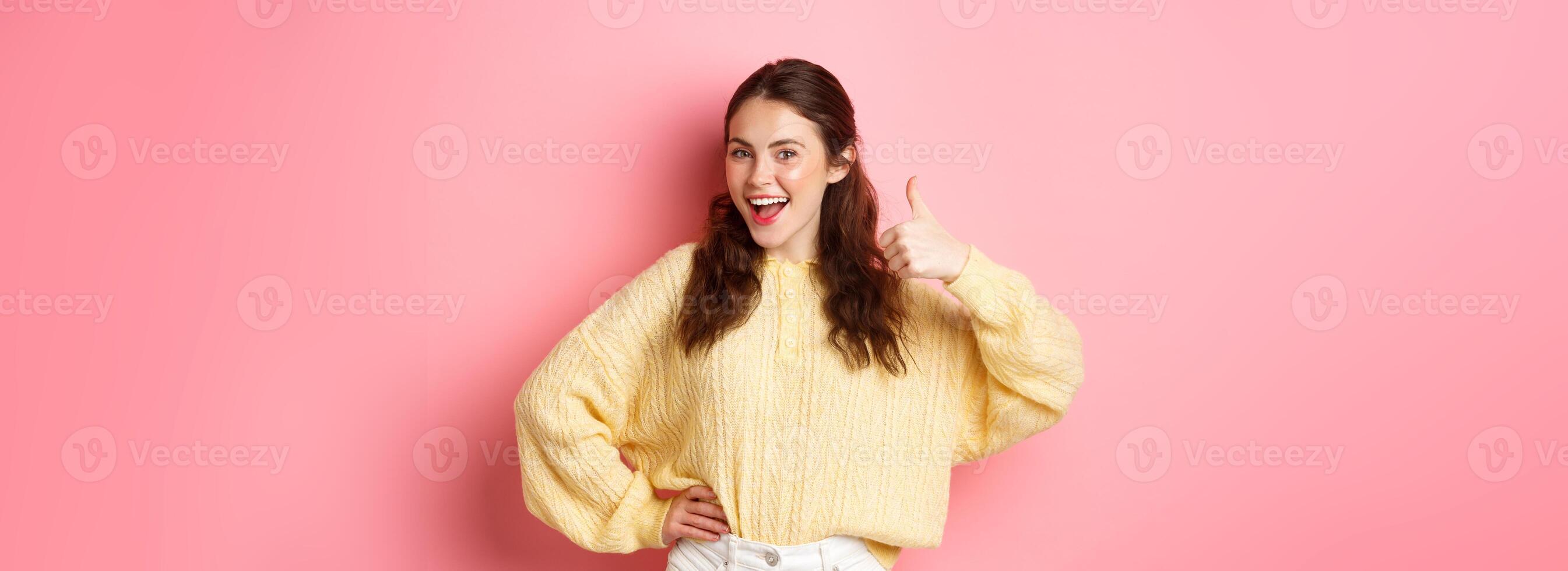 Portrait of beautiful brunette female model showing thumbs up, like your idea, give support, approve or praise something good, standing against pink background photo