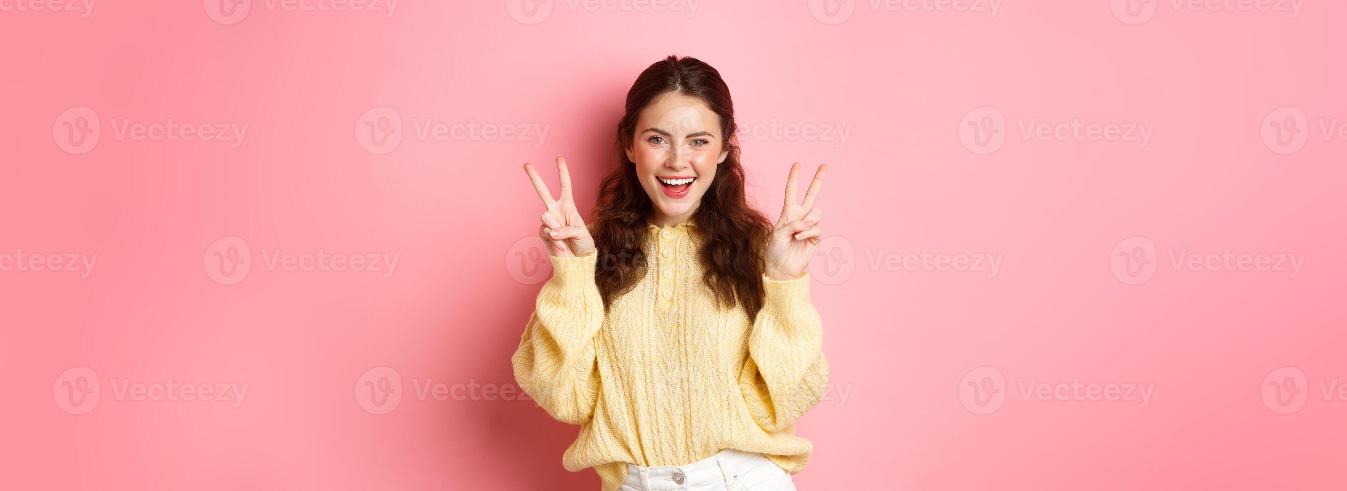 Portrait of attractive brunette female model, smiling and showing peace signs, making v-sign gesture, standing positive and happy against pink background photo
