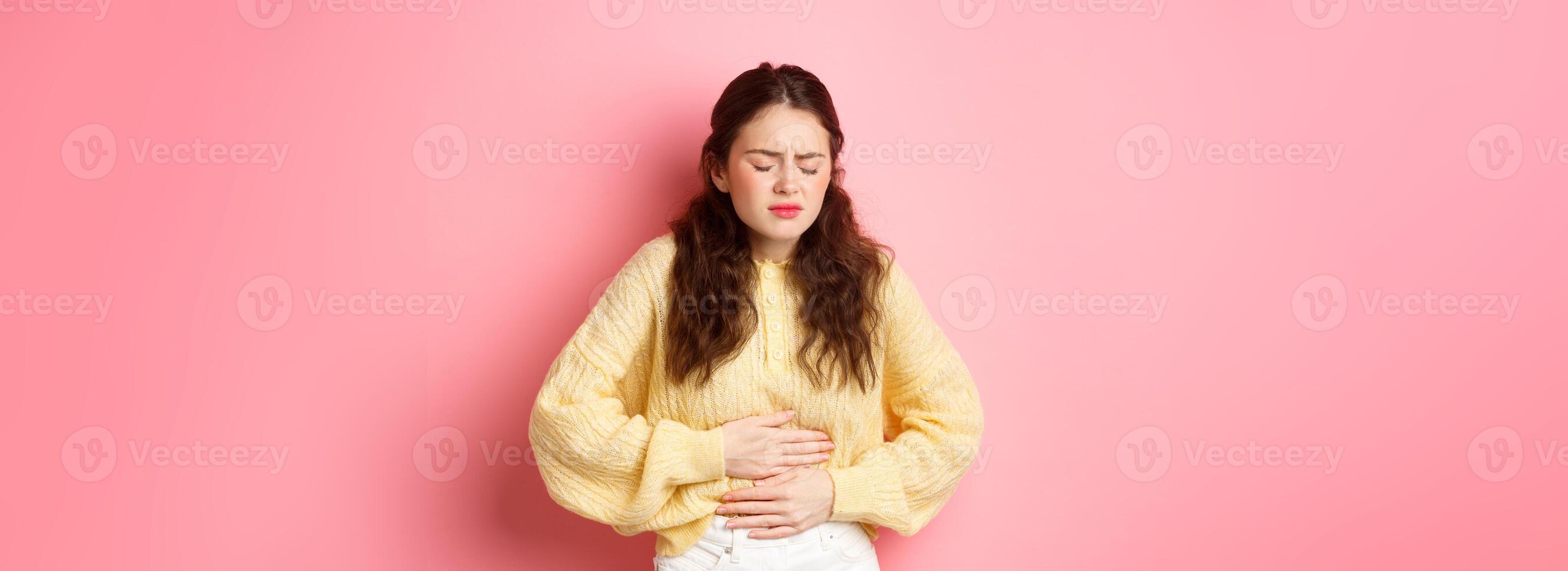Young woman with pain in stomach, holding hands on belly, feeling terrible ache, menstrual cramps, standing against pink background photo