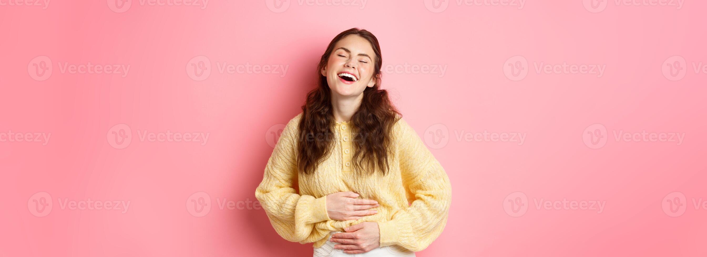 Image of carefree beautiful girl laughing out loud, touching her belly while chuckling over funny comedy, hilarious joke, standing against pink background photo