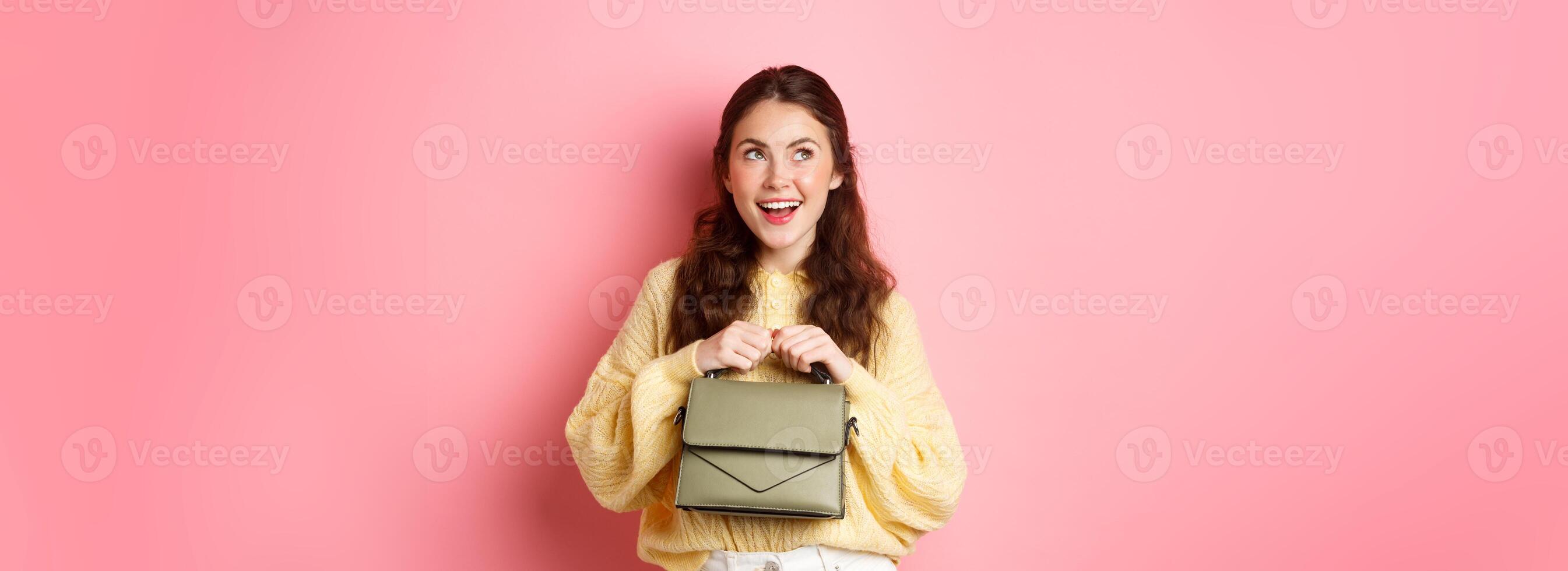 Thoughtul beautiful woman smiling, looking aside at upper left corner with pleased face, imaging something, having an idea, standing over pink background photo