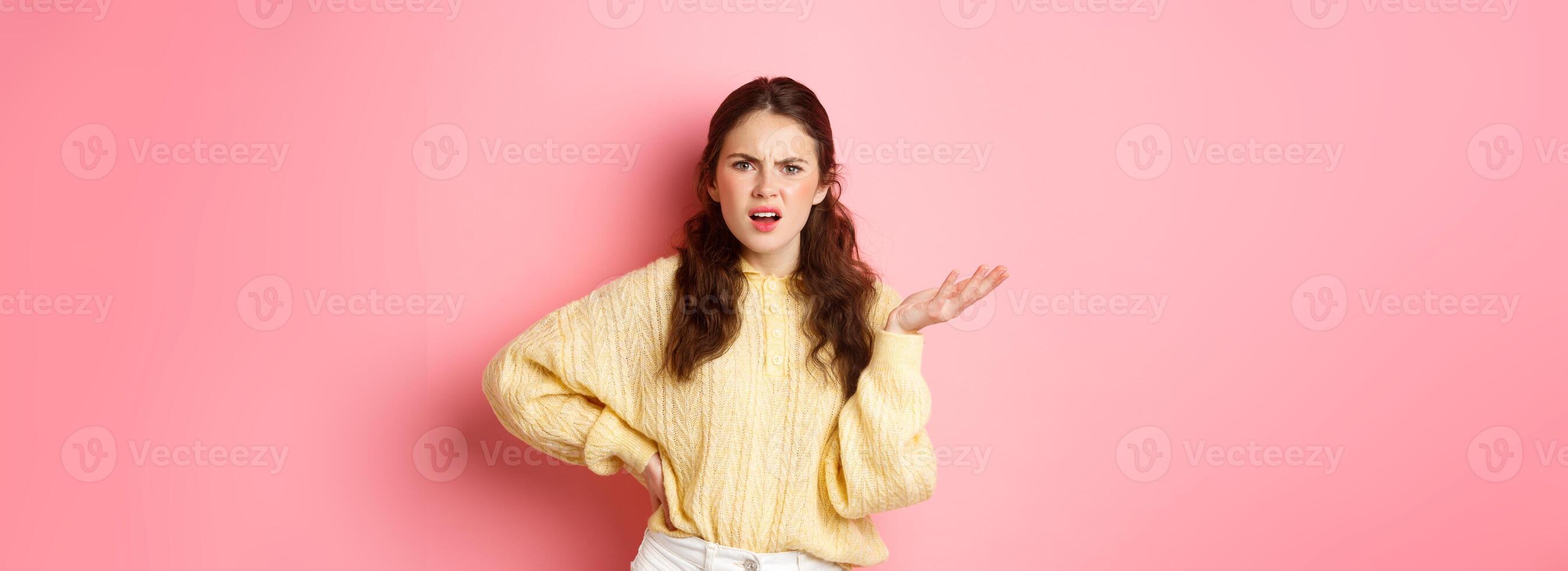 What is this, are you kidding me. Annoyed young woman complaining at something strange, staring confused, raising one hand with puzzled face, standing over pink background photo