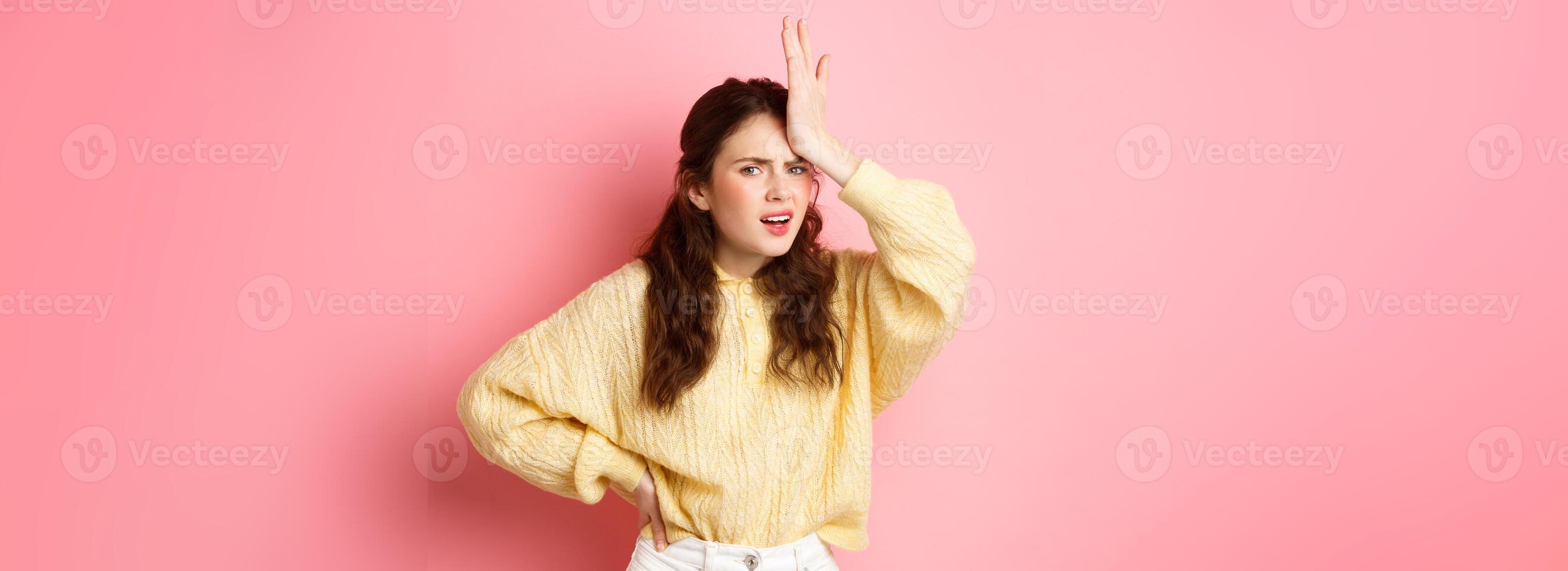 Annoyed and frustrated young woman face palm, holding hand on forehead and staring disappointed at camera, standing against pink background photo