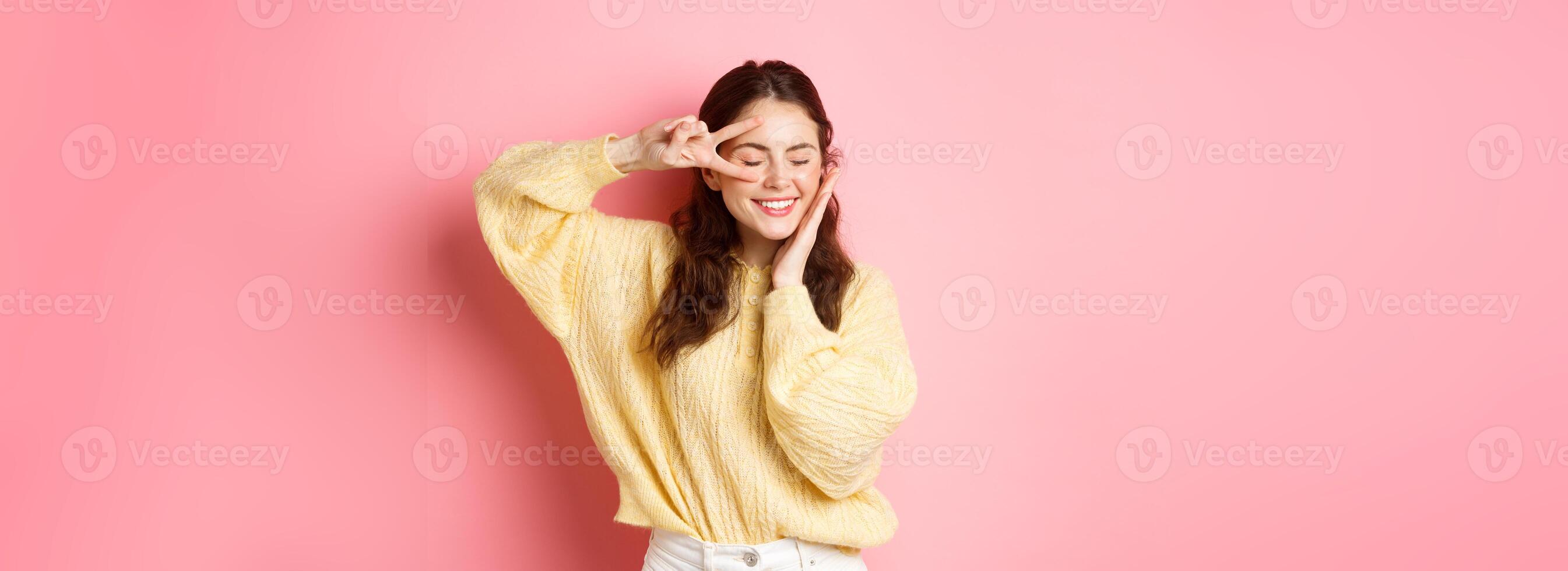 Image of beautiful brunette female model, smiling and touching her face, showing v-sign near eye, laughing happy, standing over pink background photo