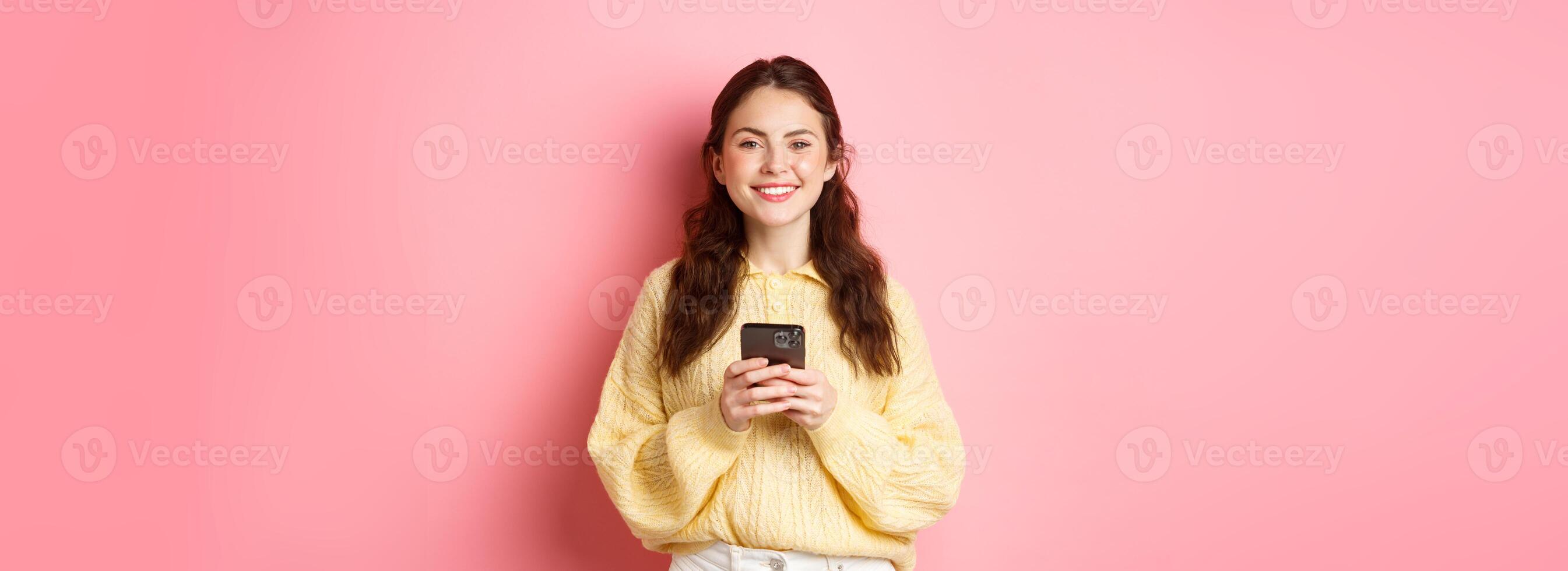 Portrait of beautiful girl holding mobile phone and smiling, shopping online, using social media app, standing against pink background in casual clothes photo