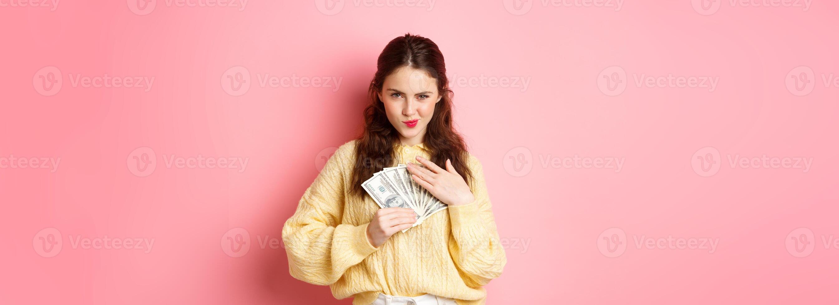 Shopping. Sassy young woman smiling and looking confident, hugging money, holding dollar bills on chest, staring thoughtful at camera, standing against pink background photo