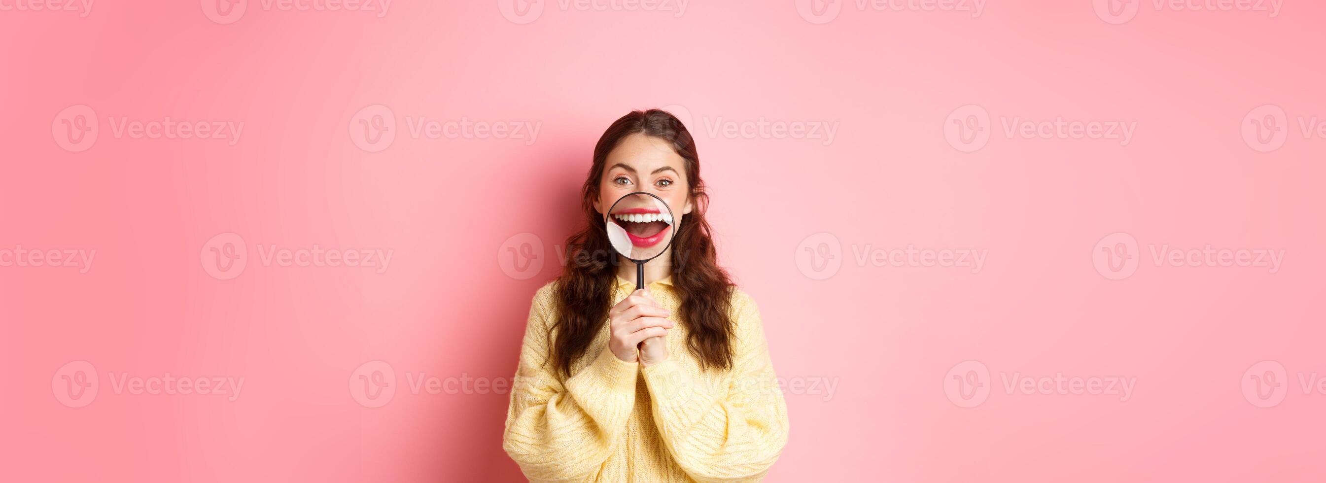 Young smiling woman showing her perfect whitened teeth smile with magnifying glass, promo of dental clinic, stomatology and healthcare concept, standing against pink background photo