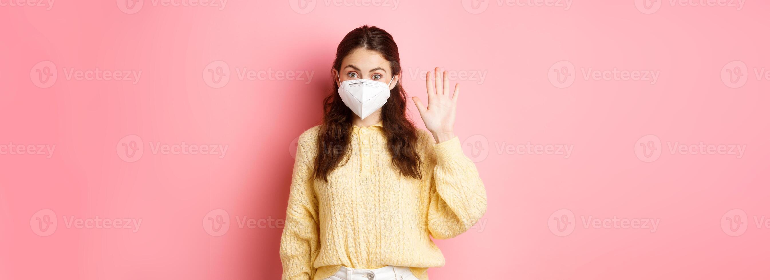 Covid-19, lockdown and pandemic concept. Young woman wearing face mask during quarantine, saying hello, waving raised hand to greet person from distance, pink background photo