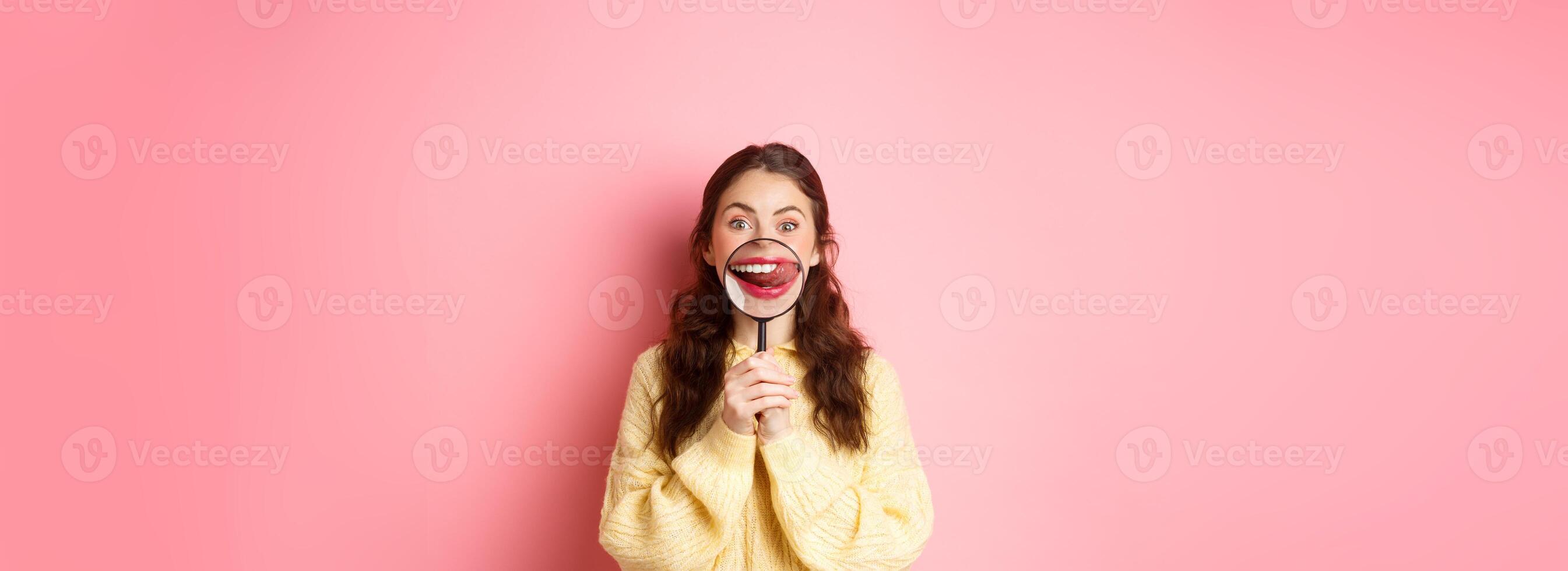 Girl looks at something tasty. Young woman shows her lips and tempted smile with magnifying glass, licking lip as staring at delicious food, standing against pink background photo