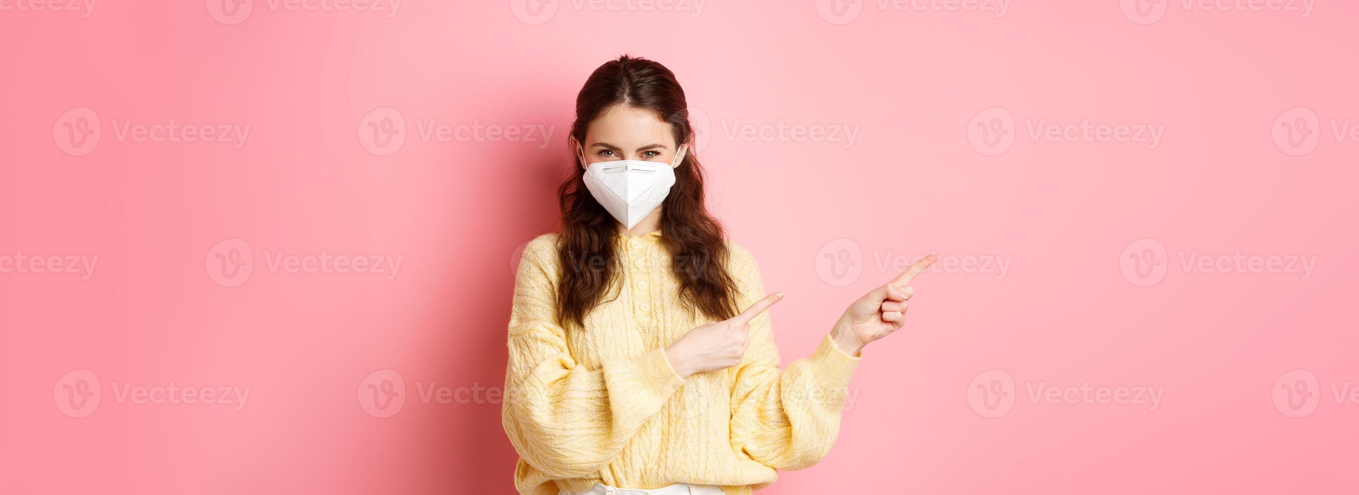 Covid, corona and social distancing concept. Coy smiling girl squinting thoughtful, pointing fingers aside at logo banner, wearing medical respirator during quarantine, pink background photo