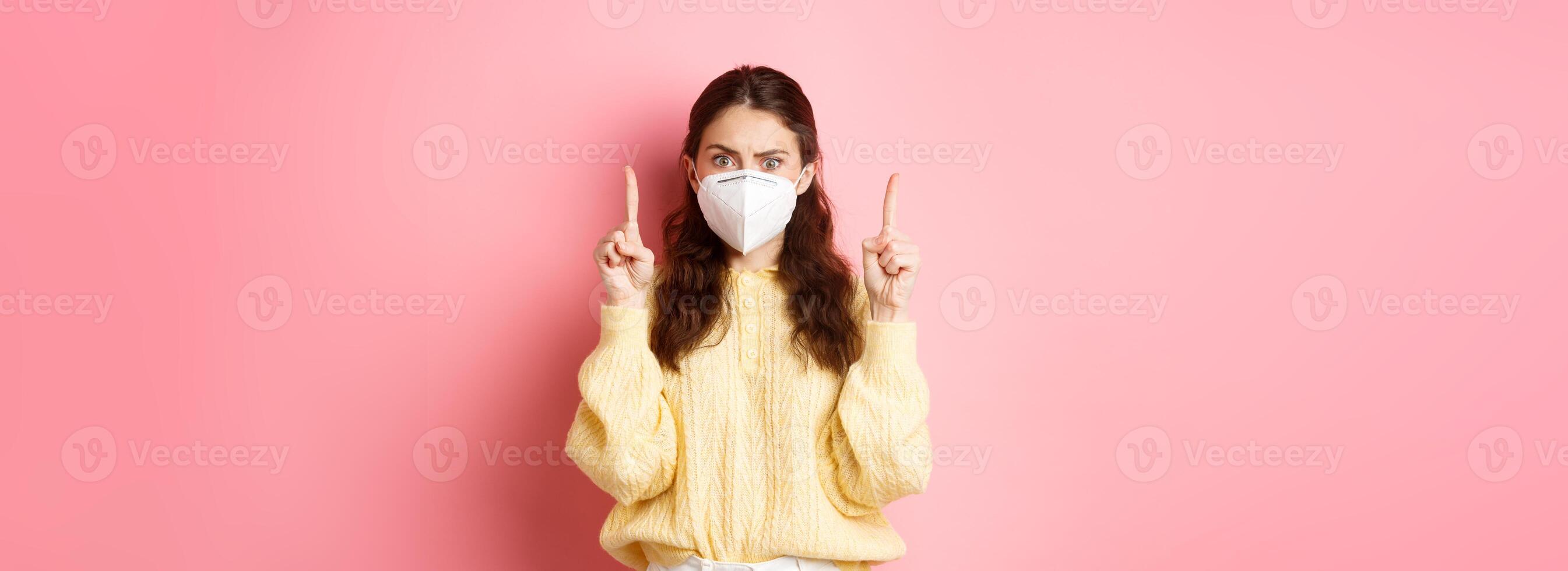 Covid, corona and social distancing concept. Confused and shocked young woman in medical respirator pointing fingers up, staring startled at camera, pink background photo