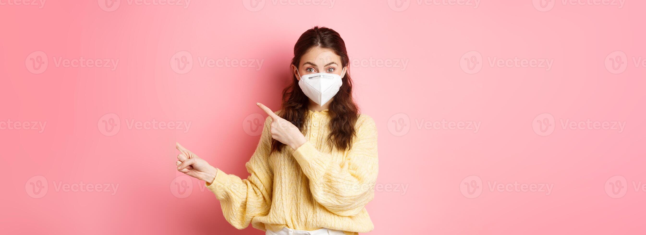 Covid, corona and social distancing concept. Excited female model in respirator shows you advertisement, points at logo aside, wears medical respirator on face, pink background photo