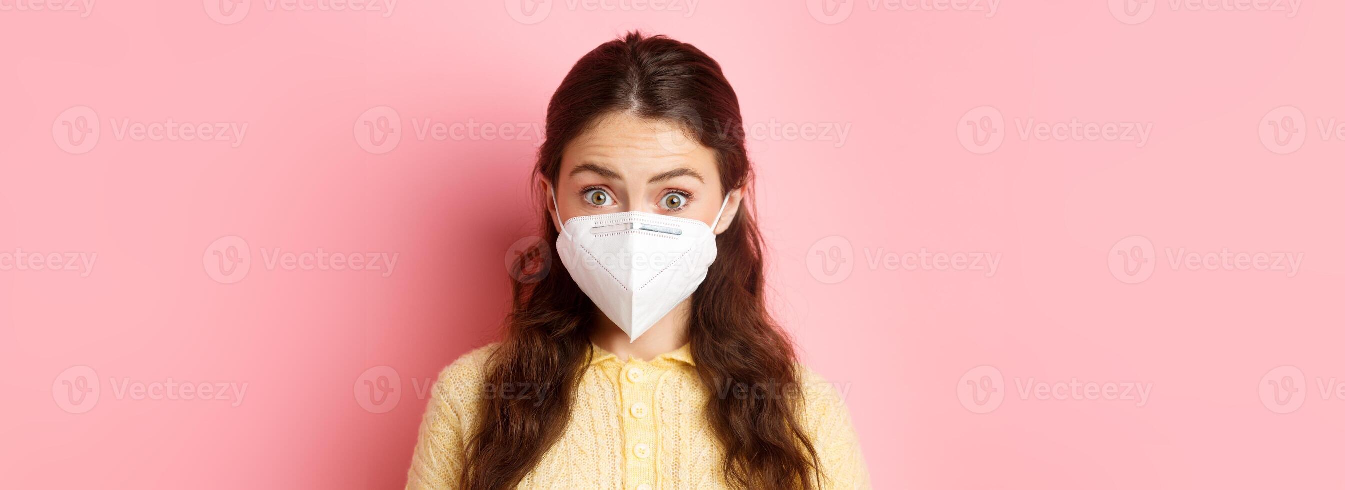 Preventive measures, health care concept. Close up portrait of woman in respirator raising eyebrows and looking surprised, hear interesting news, standing against pink background photo