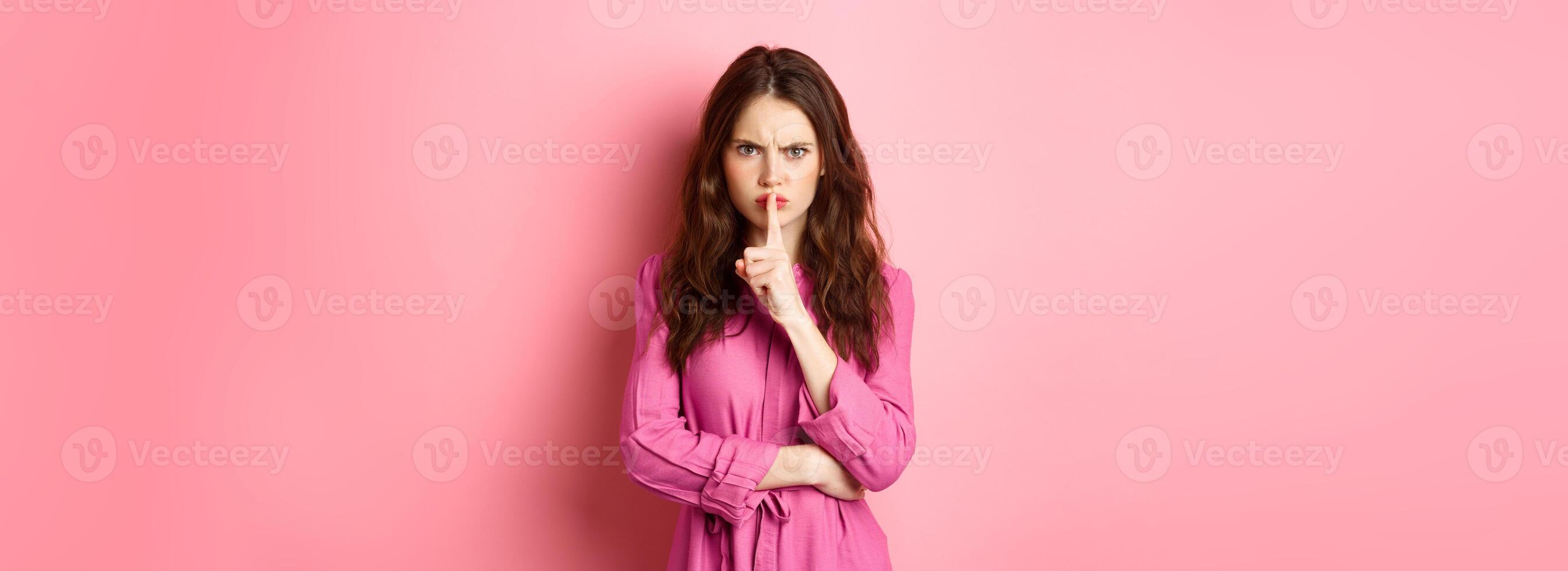 Angry girl frowning displeased, shushing at camera, press fingers to lips telling to be quiet, scolding for being loud, standing against pink background photo