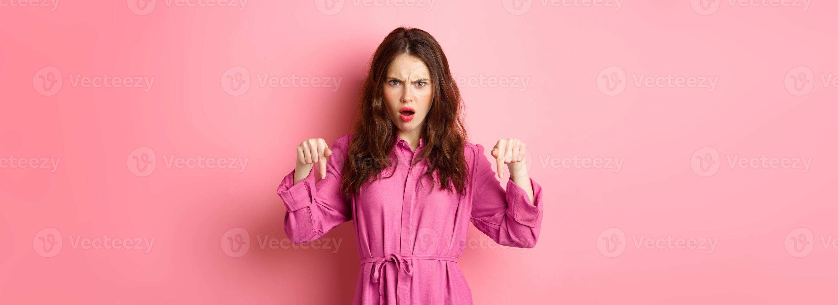 Angry and displeased young woman frowning, gasping offended, pointing fingers down, standing over pink background photo