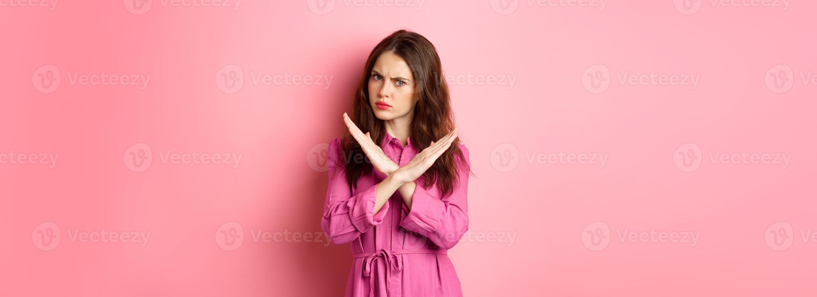 Displeased angry woman blocking offer, showing cross stop gesture, saying no and shaking head in negative reply, standing over pink background photo
