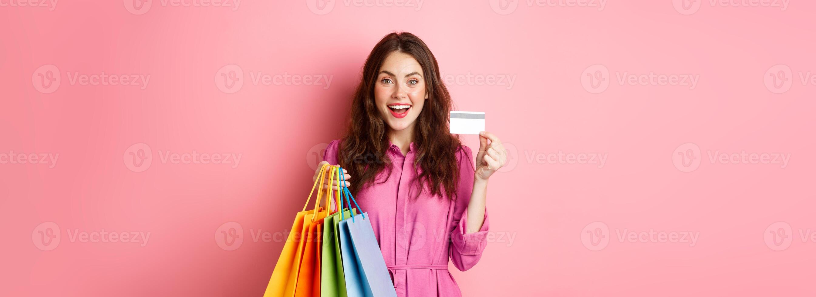 Portrait of excited happy shopaholic, woman holding shopping bags and showing plastic credit card, smiling amazed, standing against pink background photo