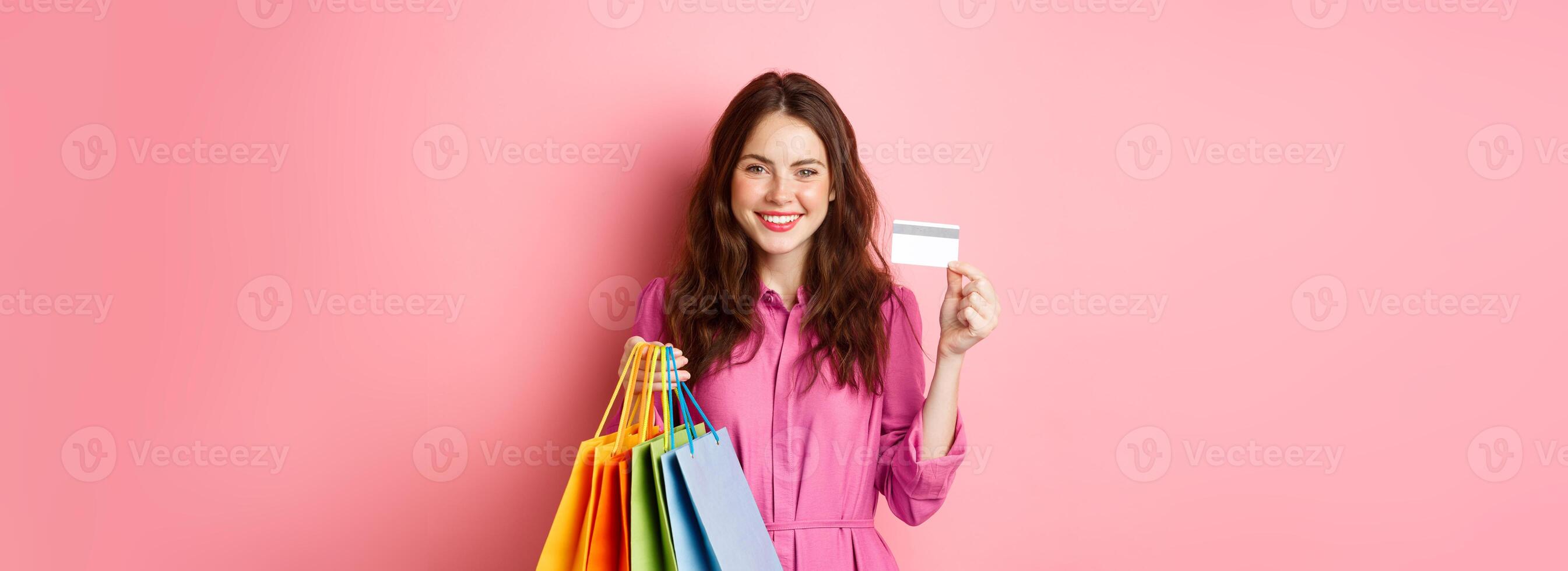 Young brunette woman holding shopping bags, showing plastic credit card and smiling, standing against pink background photo