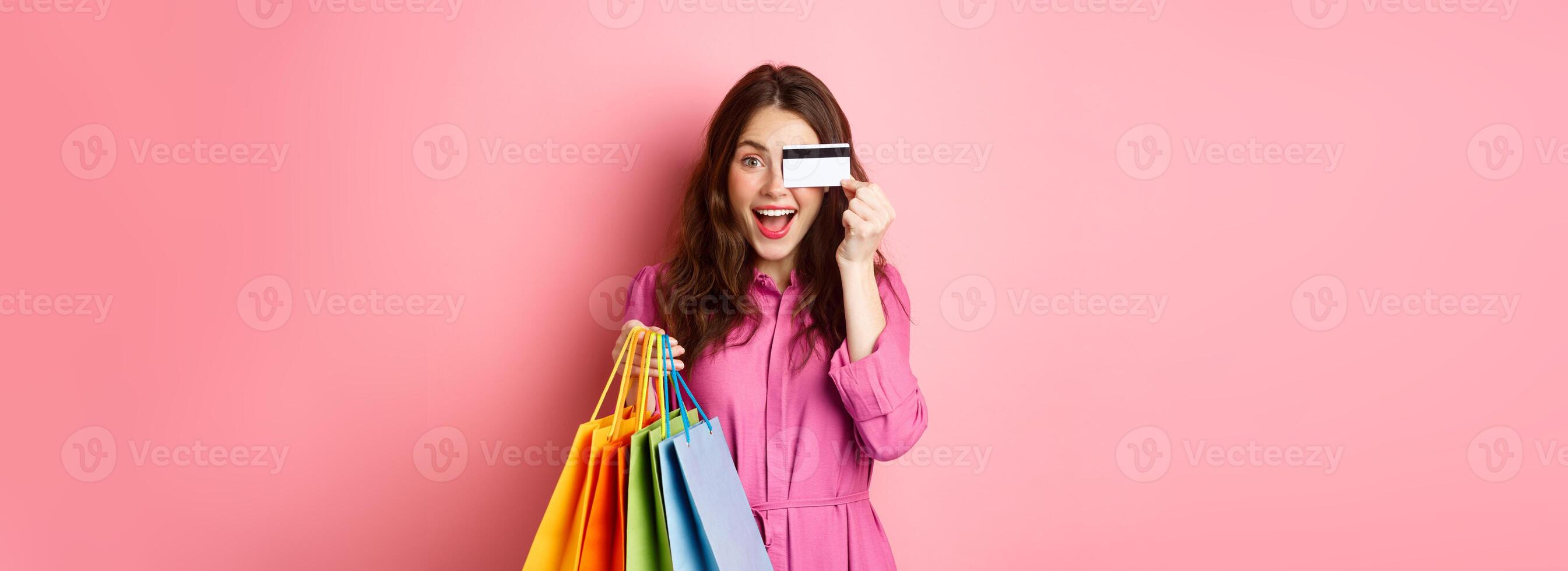 Portrait of excited happy woman holding shopping bags and showing plastic credit card, smiling amazed, standing against pink background photo
