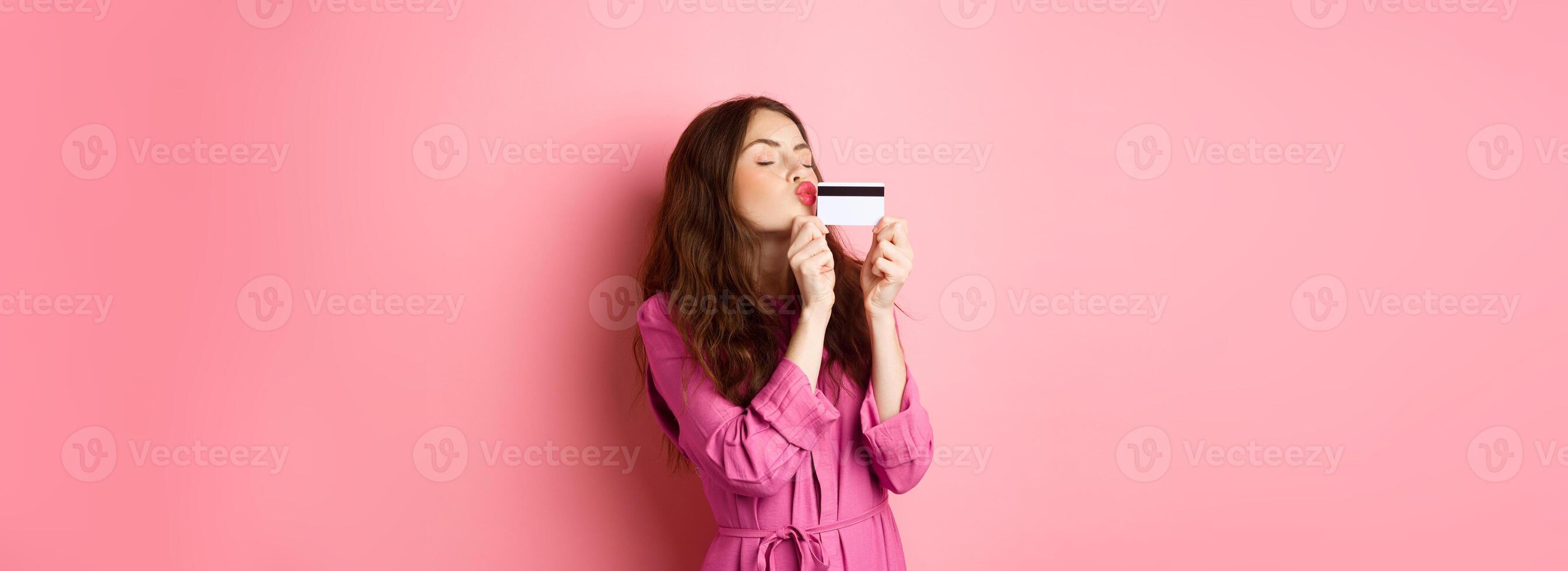 Image of young beautiful woman kissing her credit card, going on shopping, wasting money in stores, standing against pink background photo