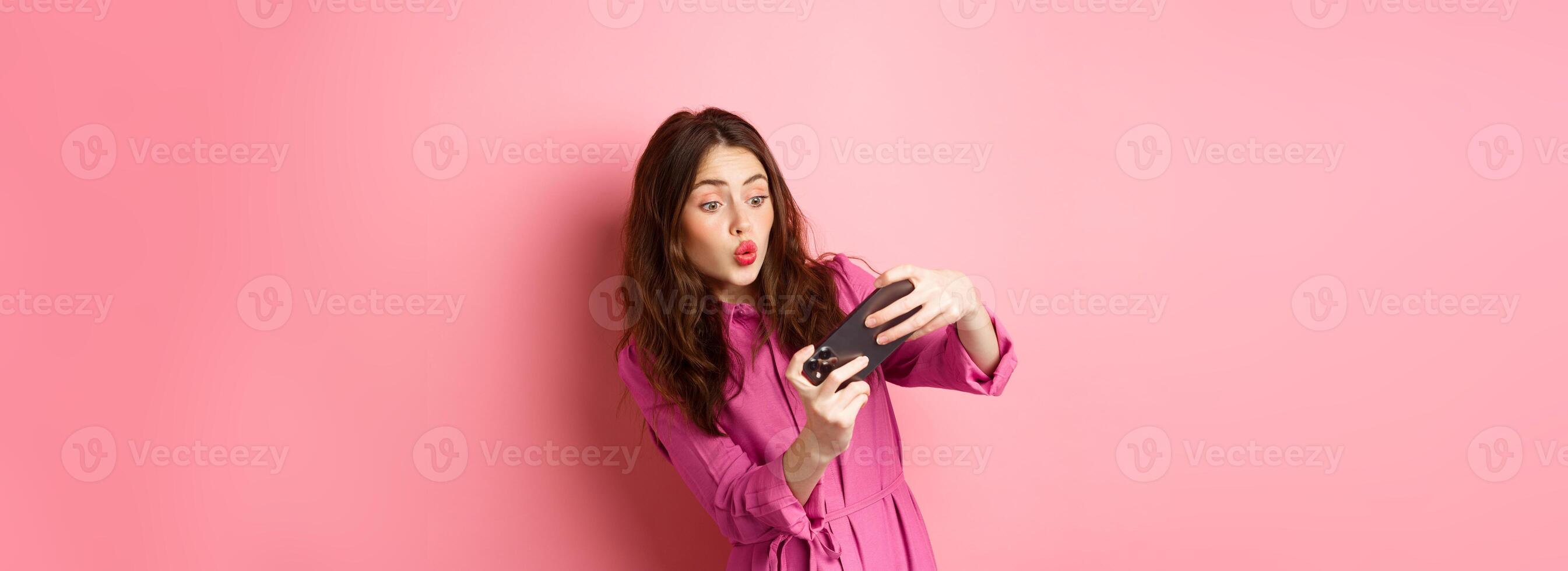 Technology concept. Carefree young woman tilt body, holding smartphone horizontally, playing game on phone, standing against pink background photo