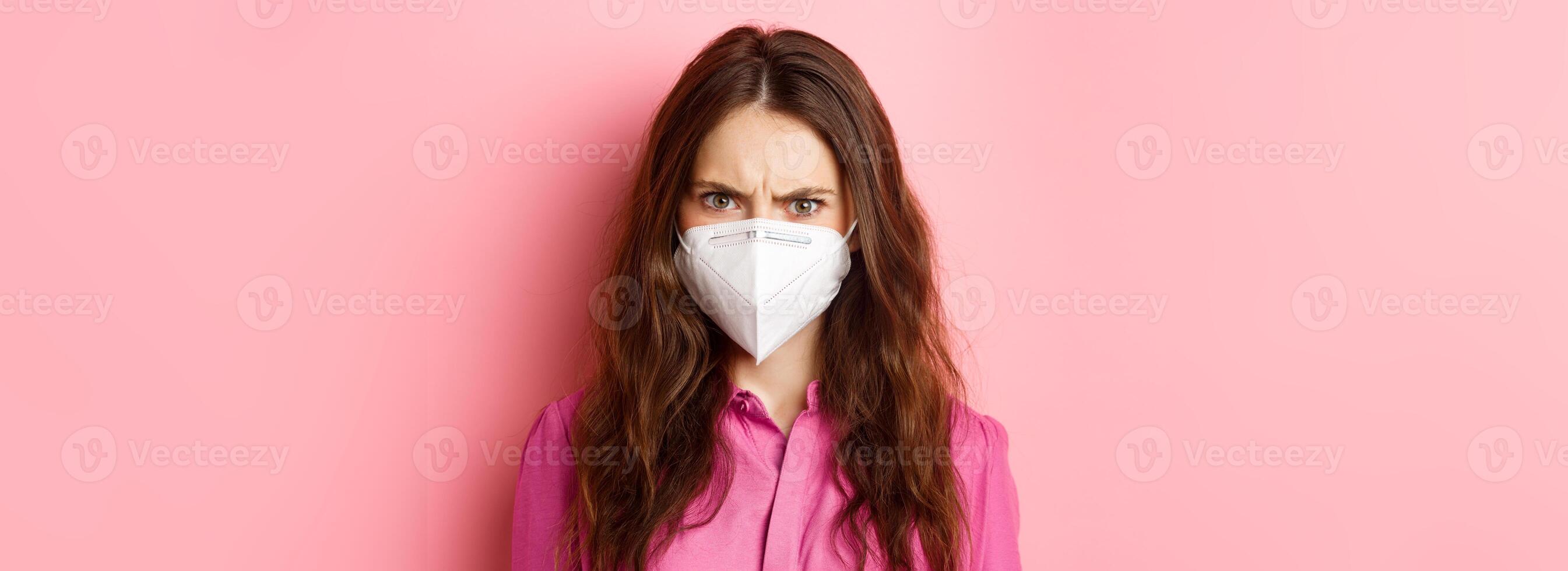 Covid-19, quarantine and social distancing concept. Close up portrait of angry female in respirator frowning, looking upset and judgemental, pink background photo