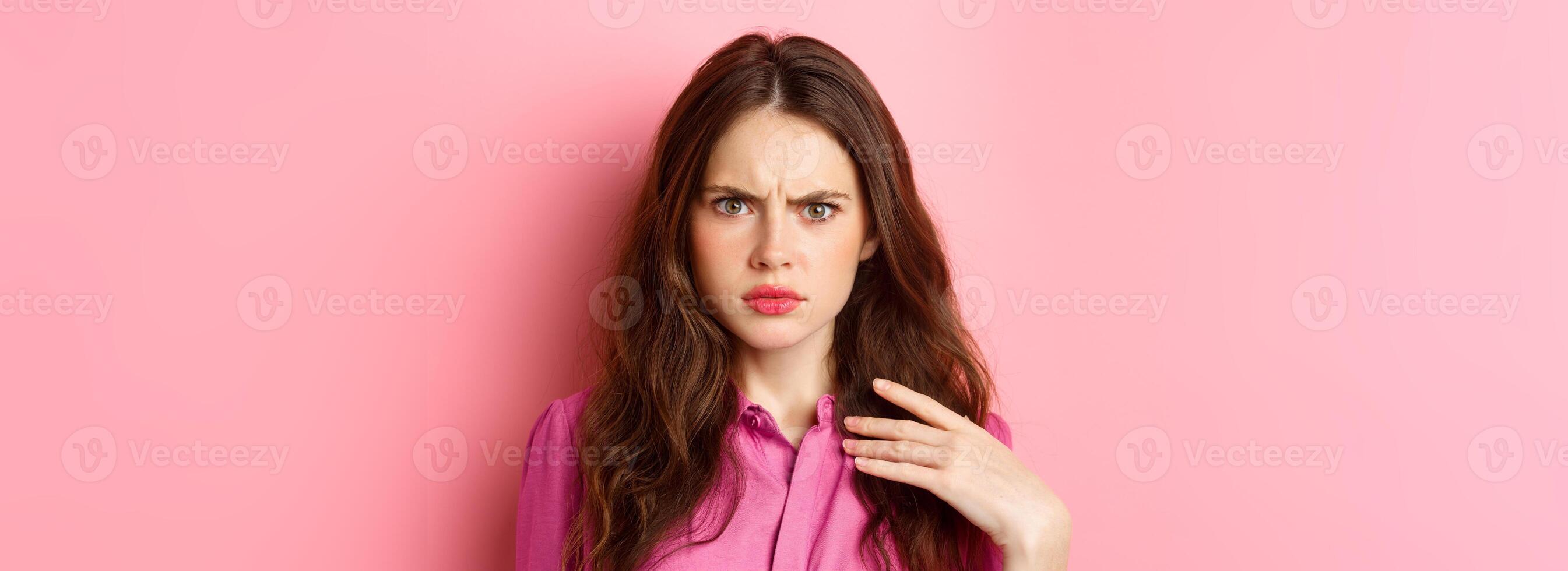 Image of arrogant young woman look with frustration and disdain, being insulted by something, frowning, holding hand on chest, pink background photo
