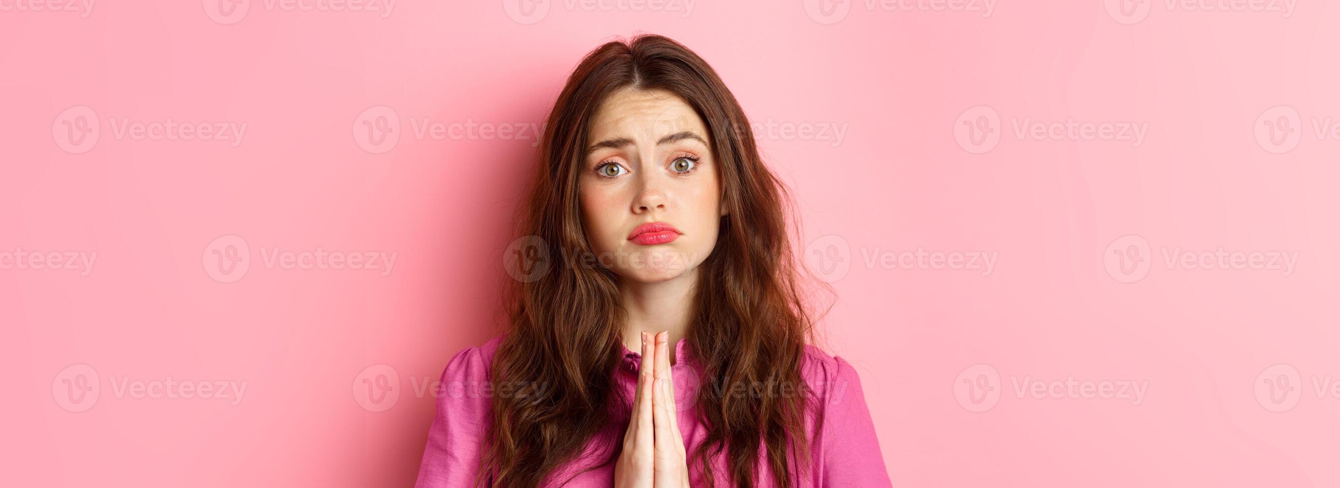 Close up of young coy girl asking for favour, holding hands in begging plead pose, making sad face, beg you, standing over pink background photo