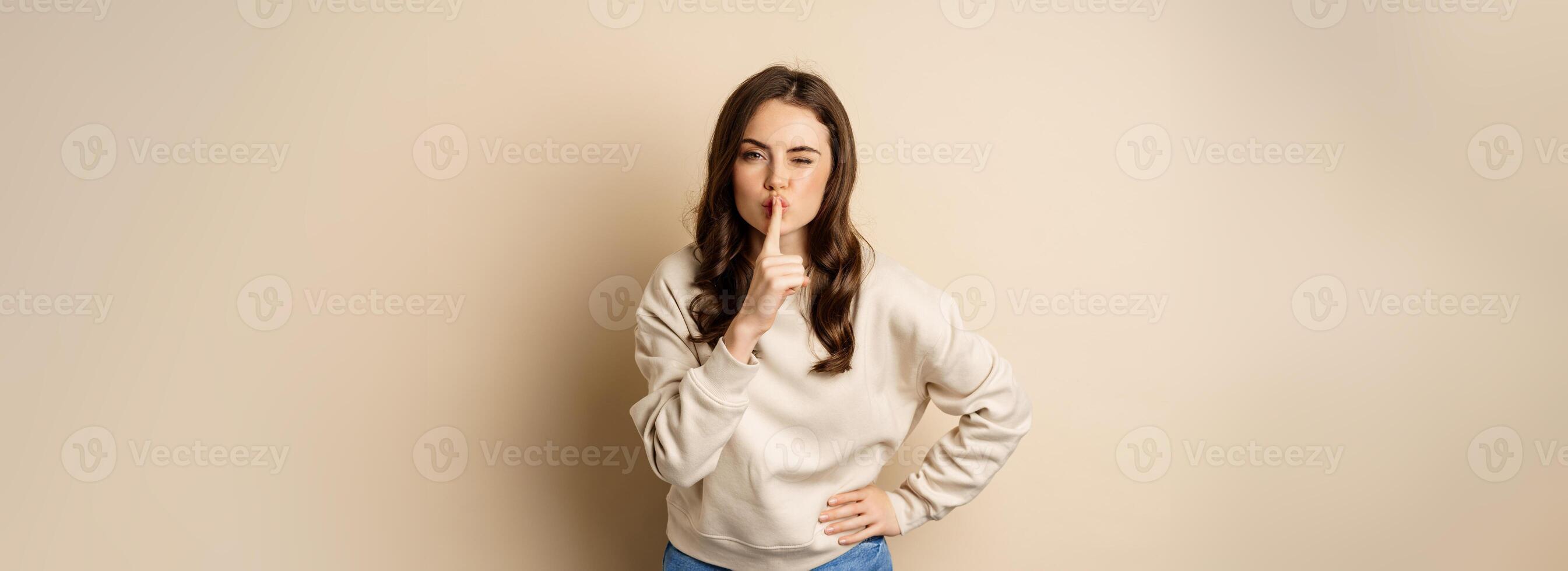 Happy coquettish girl say shh, shushing, keep secret, be quiet gesture, smiling at camera, standing over beige background photo