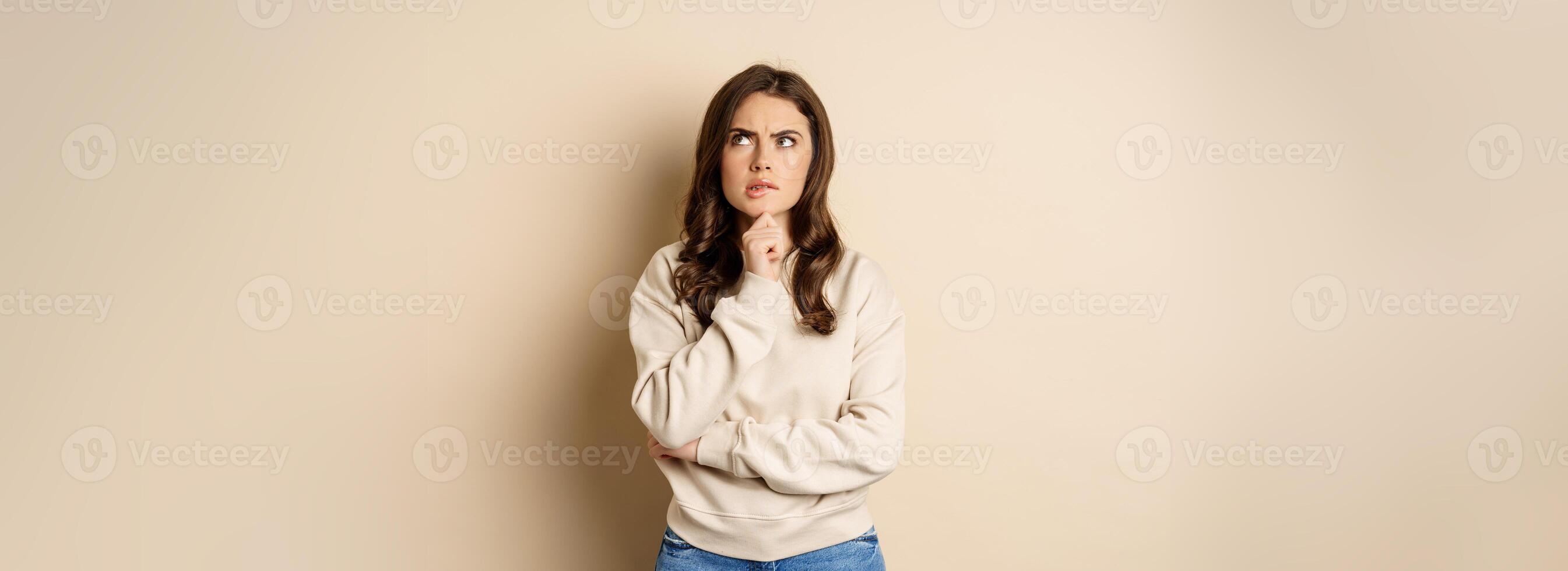 Image of beautiful woman with serious face thinking, standing over beige background photo