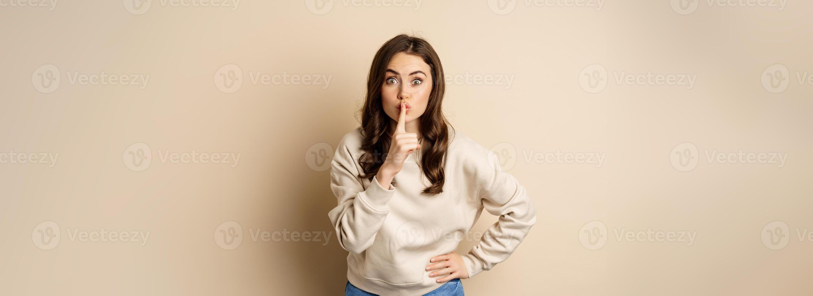 Happy coquettish girl say shh, shushing, keep secret, be quiet gesture, smiling at camera, standing over beige background photo