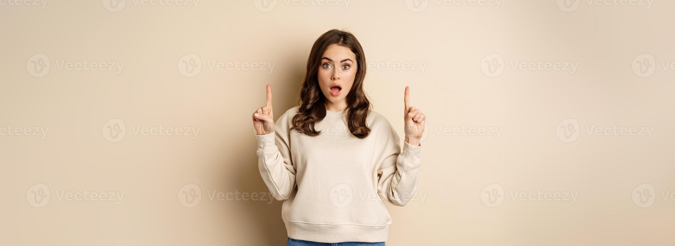 Surprised beautiful girl showing advertisement, pointing fingers on top, store sale or website link up, standing over beige background photo