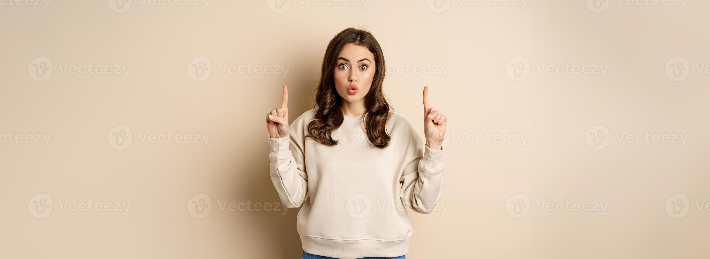 Surprised beautiful girl showing advertisement, pointing fingers on top, store sale or website link up, standing over beige background photo