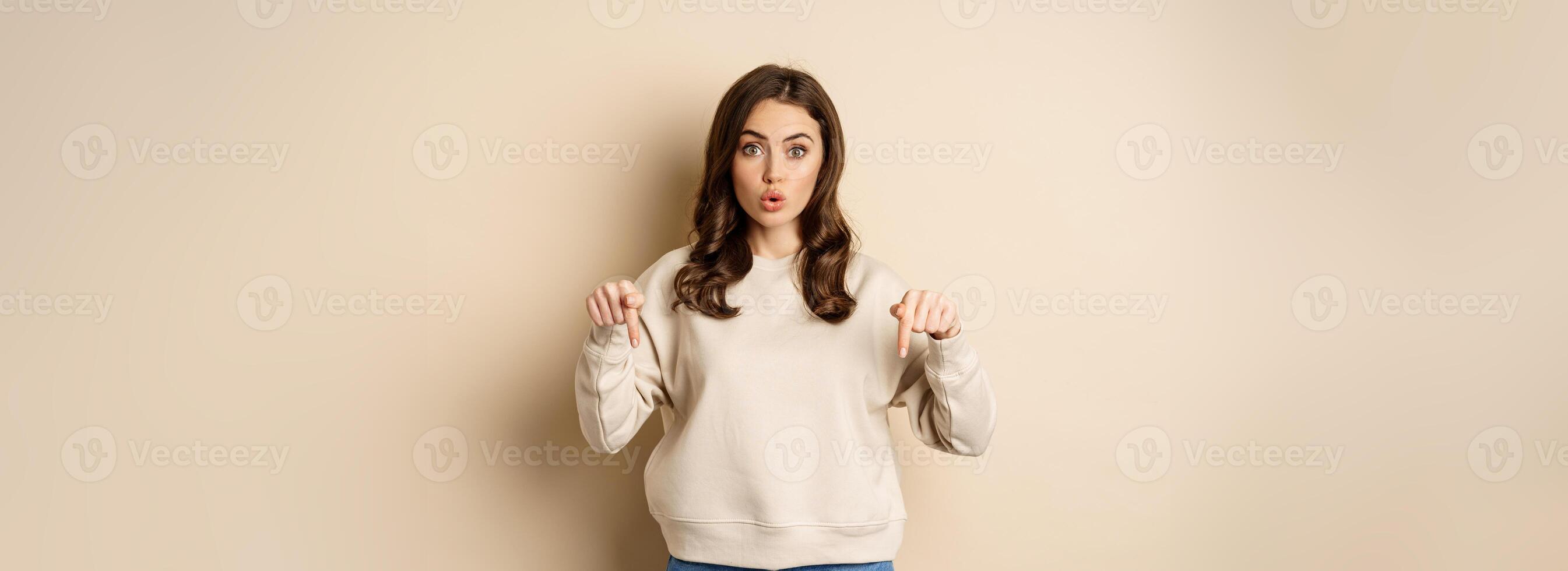Enthusiastic attractive girl pointing fingers down, showing advertisement, link or promo below, standing over beige background photo