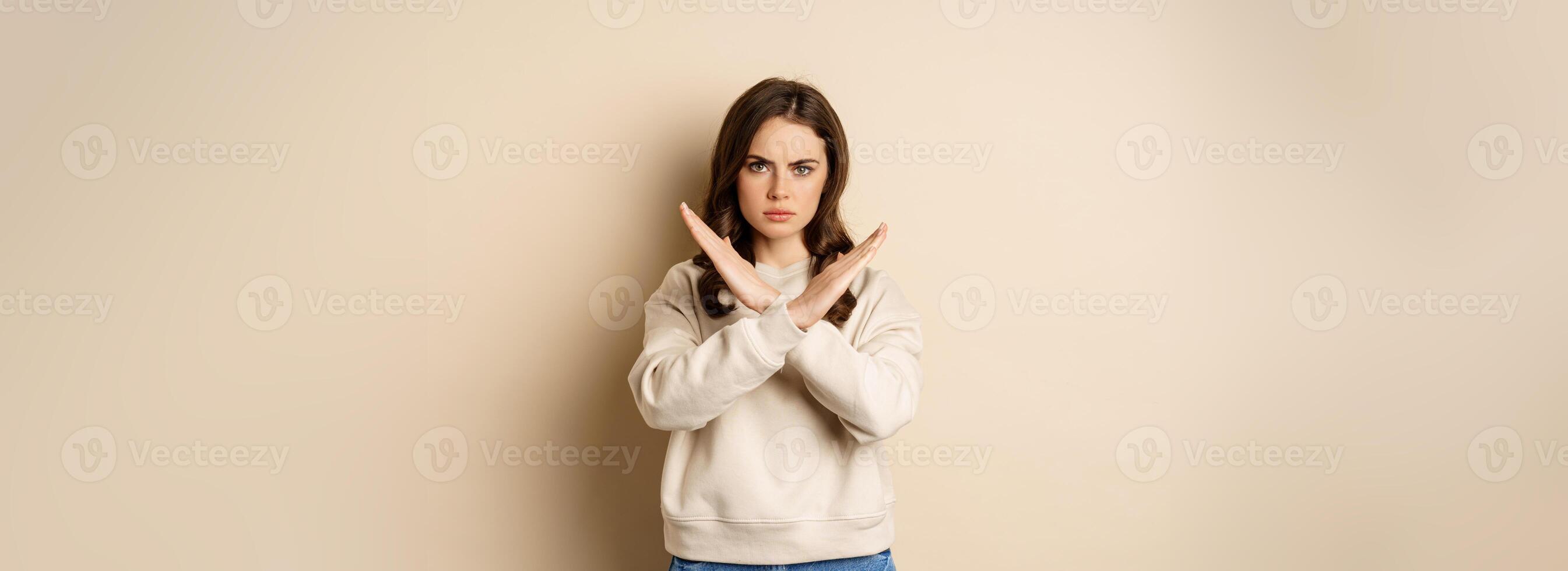 Woman showing stop, cross sign gesture and frowning angry, dislike and disapprove, saying no, rejecting and prohibiting, standing over beige background photo