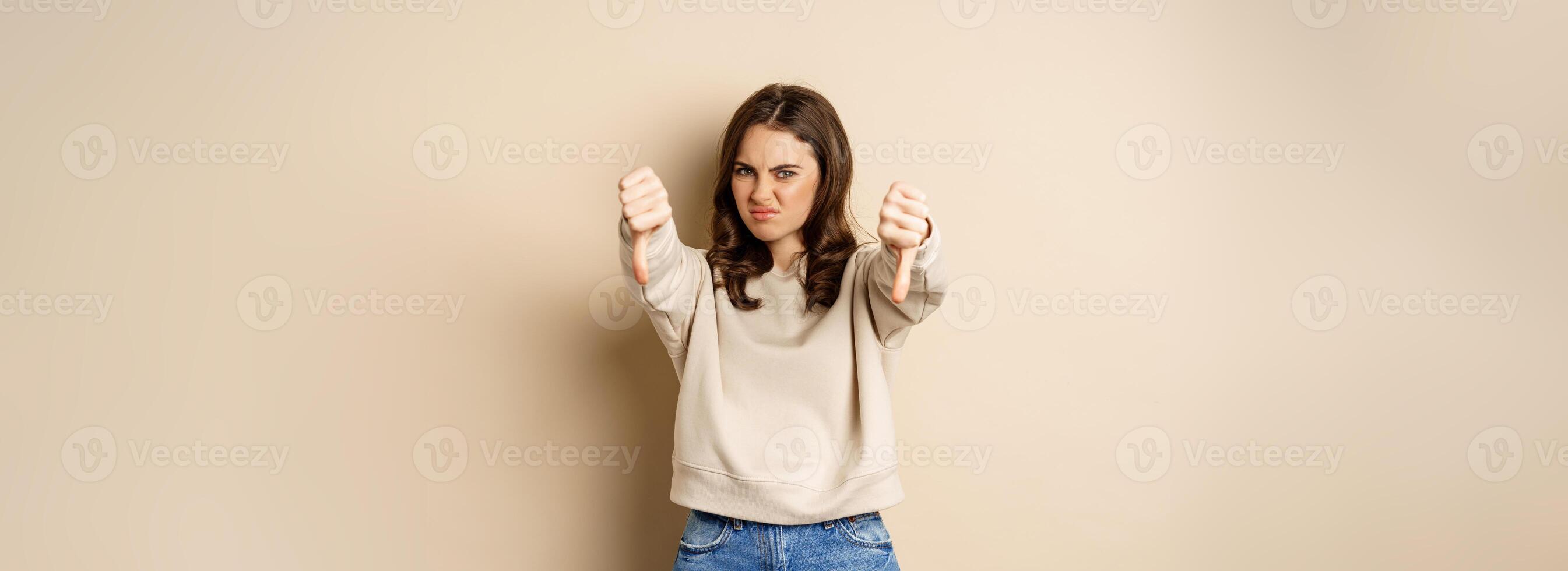 Very bad. Angry woman showing dislike, thumbs down gesture and grimacing from aversion, complaining, negative opinion feedback, beige background photo