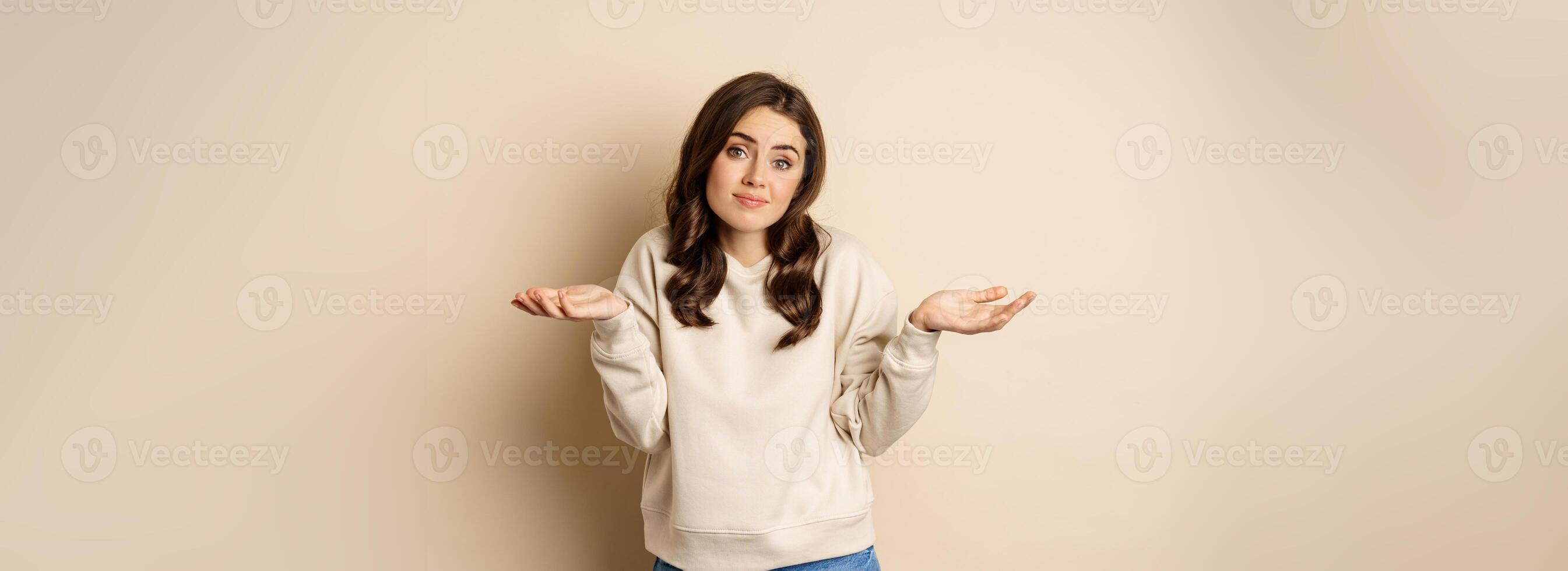 Clueless cute girl shrugging and smiling, standing over beige background photo
