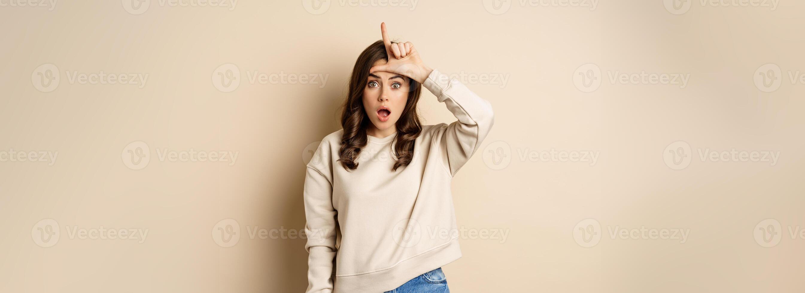 Woman showing loser sign on forehead, L word, standing over beige background photo