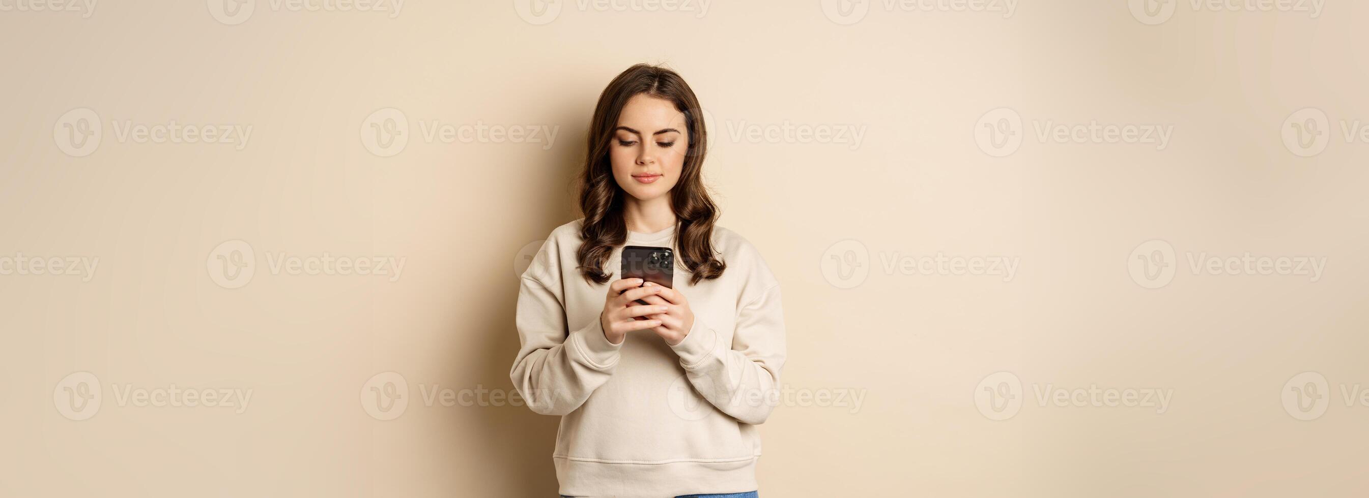 People and cellular technology. Beautiful stylish woman using mobile phone, smartphone app, smiling and looking at screen, standing over beige background photo